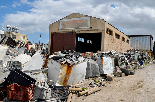 Exterior shot of a dilapidated warehouse and a cluttered scrapyard overflowing with discarded refrigerators, appliances, and various scrap metal, representing environmental waste, recycling, and industrial decay.